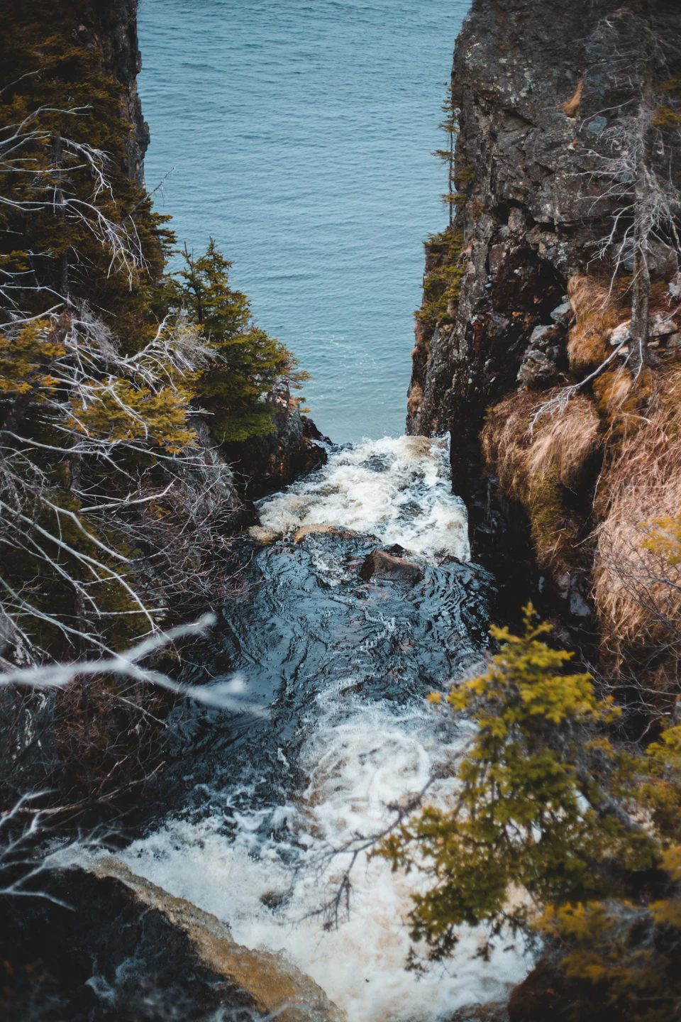 The image depicts a serene waterfall cascading down from a cliff into a river below, surrounded by lush greenery and trees. The water is a dark blue-green color, contrasting with the lighter green foliage surrounding it. The perspective of the photo is taken from above, providing an aerial view of the waterfall and its immediate surroundings.