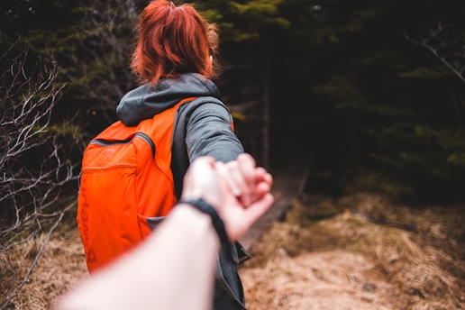 A person with red hair wearing an orange backpack is holding another person's hand, leading them along a forest path. The scene captures an outdoor adventure in a wooded area with brown grass and green trees surrounding the path.