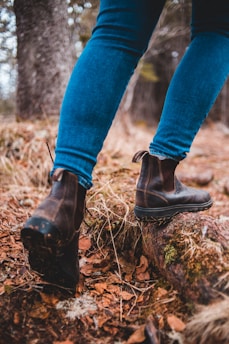 A person wearing blue denim jeans and brown leather hiking boots is stepping on a forest trail covered with fallen leaves and moss. The image focuses on the person's lower legs and feet, showcasing the rugged footwear and the natural environment.