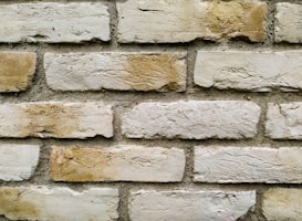 A close-up view of a brick wall with weathered, unevenly colored bricks and rough texture. The mortar between the bricks is clearly visible, giving the image an aged and rustic appearance.