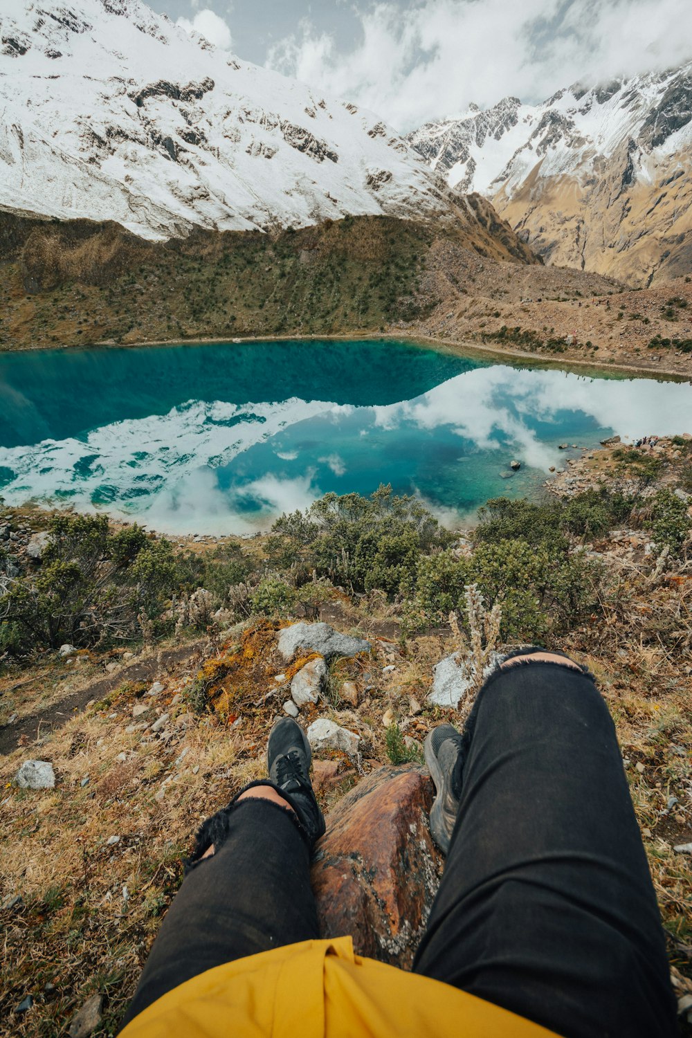 person in black pants and brown hiking shoes sitting on rock near lake during daytime