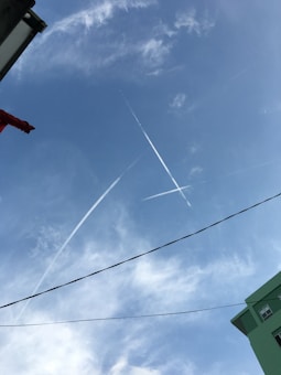 Contrails from aircraft intersect in a clear blue sky creating distinct patterns. Light clouds are scattered, and a building with green walls occupies the lower right corner. A red piece of fabric or object is partially visible at the top left.