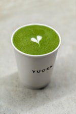 A cup filled with vibrant green matcha tea featuring a heart-shaped latte art on the surface, set against a light, neutral background. The cup has the word 'YUGEN' printed on it.