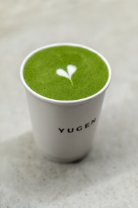 A cup filled with vibrant green matcha tea featuring a heart-shaped latte art on the surface, set against a light, neutral background. The cup has the word 'YUGEN' printed on it.