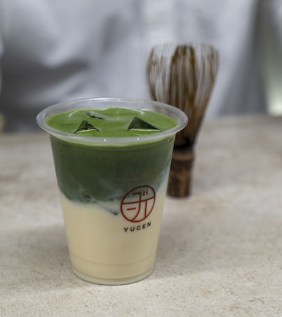 A plastic cup filled with a layered green and white beverage, likely a matcha latte, sits on a stone surface. The drink features ice cubes on top and the branding 'Yugen' on the cup. In the background, there is a bamboo whisk partially blurred.