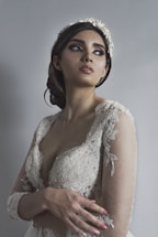 A woman wearing an elegant, lace wedding dress looks off to the side. Her hair is styled in an updo adorned with a floral headpiece. Her makeup is dramatic, emphasizing her eyes. The background is a soft gray, adding to the serene and graceful atmosphere.