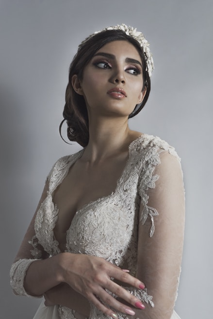 A woman wearing an elegant, lace wedding dress looks off to the side. Her hair is styled in an updo adorned with a floral headpiece. Her makeup is dramatic, emphasizing her eyes. The background is a soft gray, adding to the serene and graceful atmosphere.