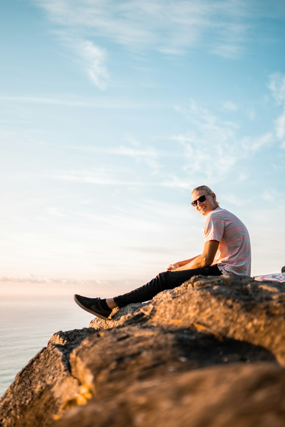 man in white t-shirt and black pants sitting on rock formation near sea during daytime