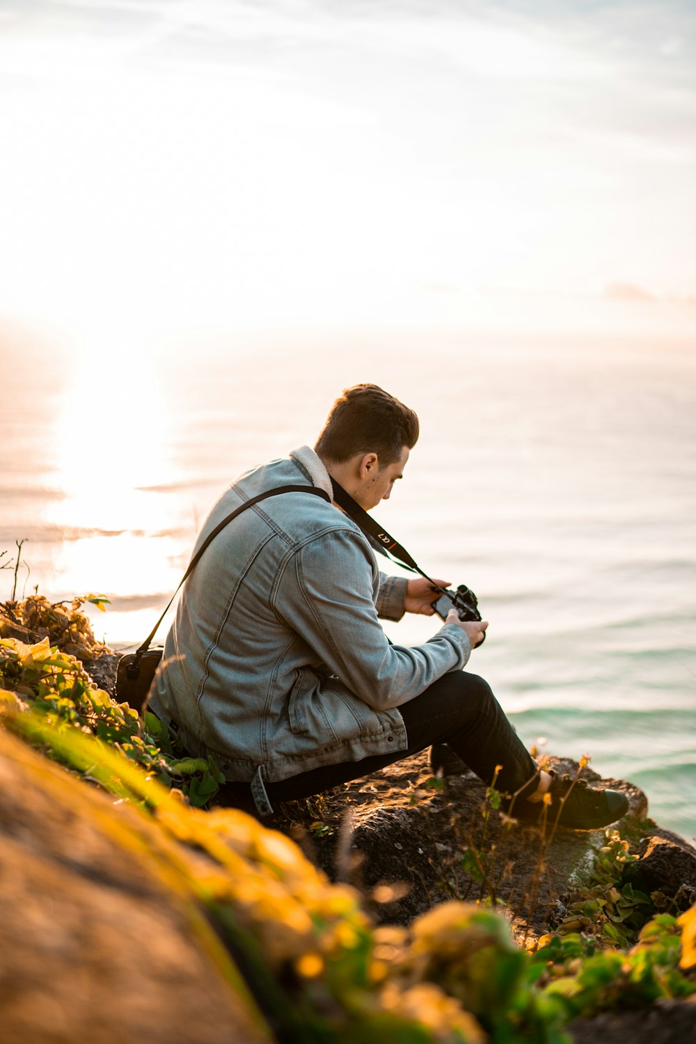 man in gray jacket sitting on rock beside body of water during daytime