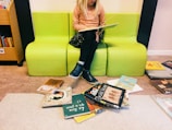 A young child with blonde hair is sitting on a bright green bench while reading a book. The child is dressed in a peach-colored sweater, black skirt, and blue shoes. Surrounding the bench, several colorful children's books are scattered on the floor. To the side, a bookshelf is partially visible with more books.
