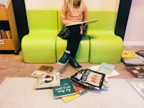 A young child with blonde hair is sitting on a bright green bench while reading a book. The child is dressed in a peach-colored sweater, black skirt, and blue shoes. Surrounding the bench, several colorful children's books are scattered on the floor. To the side, a bookshelf is partially visible with more books.