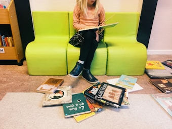 A young child with blonde hair is sitting on a bright green bench while reading a book. The child is dressed in a peach-colored sweater, black skirt, and blue shoes. Surrounding the bench, several colorful children's books are scattered on the floor. To the side, a bookshelf is partially visible with more books.