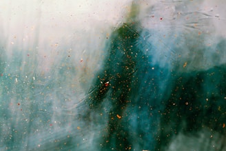 A blurry silhouette appears behind a glass surface speckled with colorful paint splatters, creating an abstract and enigmatic scene with a mix of texture and depth.