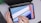 A person is holding a tablet with a stylus, creating a geometric abstract design. The tablet screen shows a digital drawing application with a user interface visible. The background includes part of a laptop keyboard and a green plant in the corner.