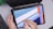A person is holding a tablet with a stylus, creating a geometric abstract design. The tablet screen shows a digital drawing application with a user interface visible. The background includes part of a laptop keyboard and a green plant in the corner.