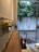 A wooden shelf is mounted on a wall inside a building, with several brass hooks lined up. Various items are placed on the shelf, including a spray bottle and two umbrellas stored below. The glass door in the background reveals a view of a concrete wall and lush green trees outside.