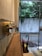 A wooden shelf is mounted on a wall inside a building, with several brass hooks lined up. Various items are placed on the shelf, including a spray bottle and two umbrellas stored below. The glass door in the background reveals a view of a concrete wall and lush green trees outside.