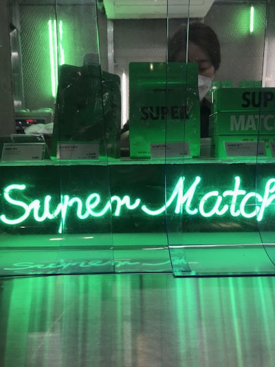 A display features neon green signage with the words 'Super Match' illuminated brightly. Behind the glass, several products in green packaging are visible, and a person wearing a mask can be seen in the background. The overall ambiance is modern with reflective surfaces.