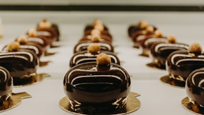 Rows of glossy, chocolate-coated pastries are neatly arranged on a reflective surface, each topped with a small garnish. The pastries are uniform in shape and are set in a well-lit display, highlighting their smooth and luxurious finish.