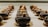 Rows of glossy, chocolate-coated pastries are neatly arranged on a reflective surface, each topped with a small garnish. The pastries are uniform in shape and are set in a well-lit display, highlighting their smooth and luxurious finish.