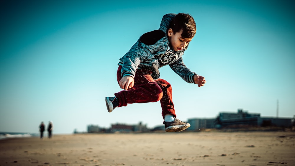 boy in blue jacket and red pants jumping on brown field during daytime