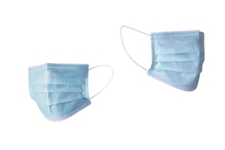 Two disposable medical face masks in light blue are suspended on a plain white background. Each mask features pleated layers and elastic ear loops.