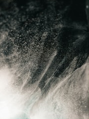 A dynamic swirl of fine mist or dust particles creates an abstract pattern against a dark background, with light creating a shimmering effect.