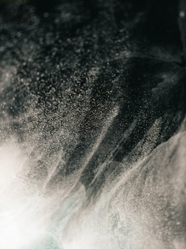 A dynamic swirl of fine mist or dust particles creates an abstract pattern against a dark background, with light creating a shimmering effect.