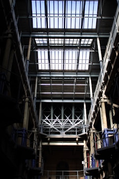 A large industrial interior with a high, glass-panelled roof allowing natural light to flood in. Metal beams and columns frame the view, creating a sense of symmetry and grandeur. The structure is robust, with intricate trusses and multiple levels visible, evoking a historical industrial architecture feel.