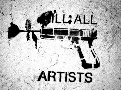 A black stencil graffiti on a rough, textured wall featuring a gun with a flower coming out of the barrel. Above the gun are the words 'ILL ALL' and below it the word 'ARTISTS' is visible.