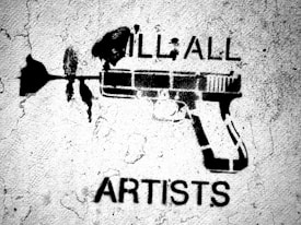 A black stencil graffiti on a rough, textured wall featuring a gun with a flower coming out of the barrel. Above the gun are the words 'ILL ALL' and below it the word 'ARTISTS' is visible.