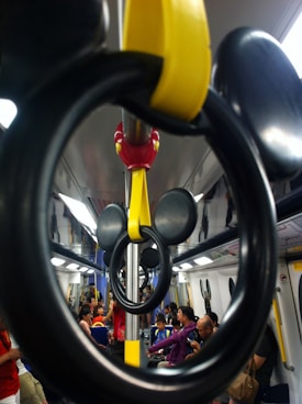 A train interior featuring handrails designed to resemble mouse ears, with passengers seated and standing, some interacting with electronic devices. The carriage is brightly lit with a mix of blue, yellow, and black colors, and people are casually dressed.