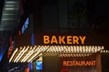 A neon sign reading 'BAKERY' in bright orange letters is prominently displayed above a shop entrance at night. Below the sign, a line of small lights adds further illumination. The word 'RESTAURANT' is also visible in neon, along with the establishment year sign 'EST. 1950' on the side.