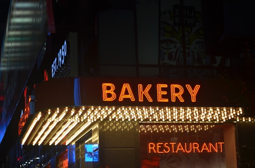 A neon sign reading 'BAKERY' in bright orange letters is prominently displayed above a shop entrance at night. Below the sign, a line of small lights adds further illumination. The word 'RESTAURANT' is also visible in neon, along with the establishment year sign 'EST. 1950' on the side.