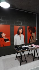 The image features three large portraits, each depicting a woman against a vibrant red background. In front of the portraits, a wooden table displays various neatly arranged clothing items, including hats and folded shirts. The setting appears to be indoors, with a dimly lit overhead lamp and a dark, industrial wall in the background.