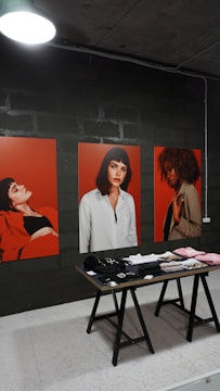 The image features three large portraits, each depicting a woman against a vibrant red background. In front of the portraits, a wooden table displays various neatly arranged clothing items, including hats and folded shirts. The setting appears to be indoors, with a dimly lit overhead lamp and a dark, industrial wall in the background.