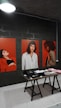 The image features three large portraits, each depicting a woman against a vibrant red background. In front of the portraits, a wooden table displays various neatly arranged clothing items, including hats and folded shirts. The setting appears to be indoors, with a dimly lit overhead lamp and a dark, industrial wall in the background.