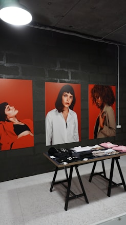 The image features three large portraits, each depicting a woman against a vibrant red background. In front of the portraits, a wooden table displays various neatly arranged clothing items, including hats and folded shirts. The setting appears to be indoors, with a dimly lit overhead lamp and a dark, industrial wall in the background.