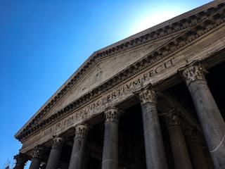 The image features the facade of a classical ancient temple with large stone columns and a detailed pediment. Latin inscriptions are visible on the frieze, and the structure is set against a clear, bright blue sky.