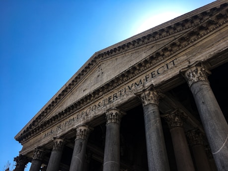 The image features the facade of a classical ancient temple with large stone columns and a detailed pediment. Latin inscriptions are visible on the frieze, and the structure is set against a clear, bright blue sky.