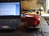 A cozy café scene with a laptop on the left side of the table and a red coffee cup on a saucer in the foreground. Several people are seated in the background, engaging in conversation and reading. The café is dimly lit, with hanging lights and a relaxed ambiance. Potted flowers can be seen on some of the tables, adding a touch of color to the setting.