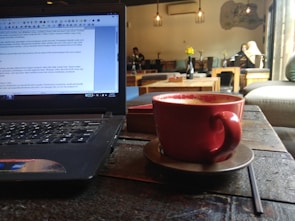 A cozy café scene with a laptop on the left side of the table and a red coffee cup on a saucer in the foreground. Several people are seated in the background, engaging in conversation and reading. The café is dimly lit, with hanging lights and a relaxed ambiance. Potted flowers can be seen on some of the tables, adding a touch of color to the setting.