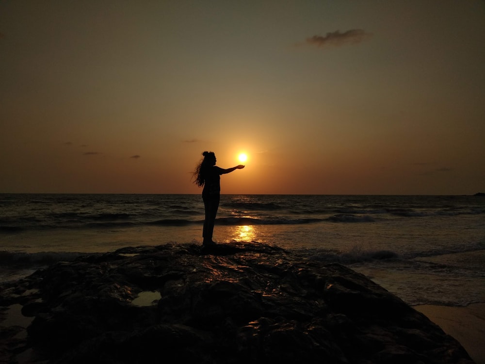 silhouette of woman standing on rocky shore during sunset