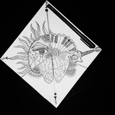 A detailed black and white drawing incorporating a sun with a stylized eye, elements of a compass, a brain, and a guitar. The artwork includes sun rays transitioning into piano keys and ornate patterns radiating from the central focus. A pen is placed near the top edge.