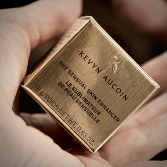 A hand holding a small, metallic gold box labeled 'Kevyn Aucoin The Sensual Skin Enhancer'. The text is in both English and French, noting the weight as 18g or 0.63 oz.