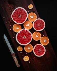 Slices of bright red grapefruits and orange-colored citrus fruits are artfully arranged on a dark wooden board. A rustic knife with a patina-riddled blade rests alongside the fruit, adding a touch of rustic charm. Scattered pomegranate seeds accent the composition with additional color contrast.