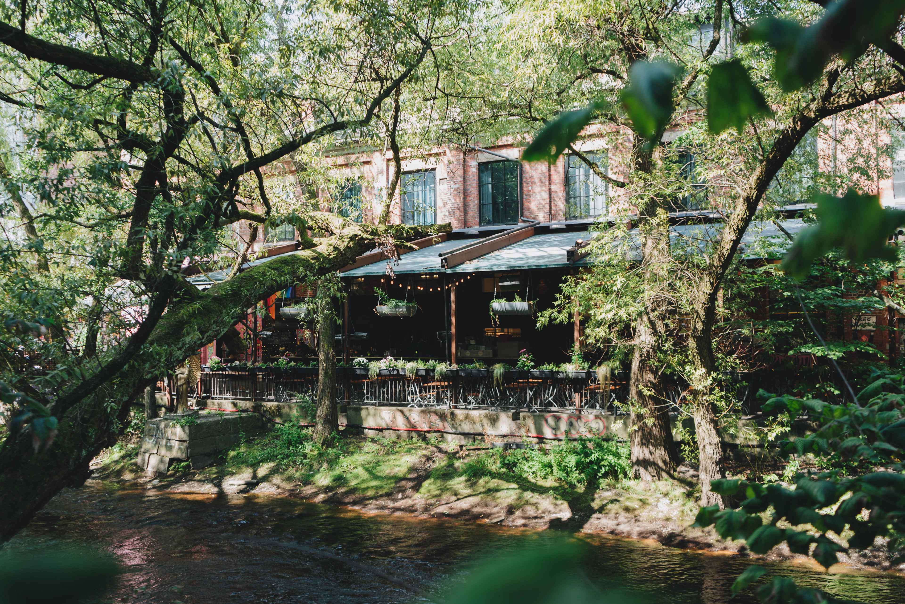An old brick mill building on the Akerselva river in Oslo, its riverside terrace strung with festoon lights and set with tables, framed by overhanging trees in full summer leaf above the running water.