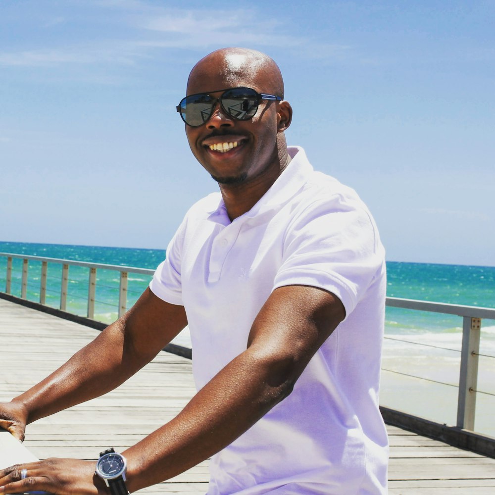 man in white polo shirt wearing sunglasses sitting on brown wooden dock during daytime