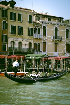 A picturesque canal scene with a gondolier navigating a sleek black gondola through the water. Passengers, leisurely seated under a shaded canopy, enjoy the view of historic, pastel-colored buildings along the water's edge. The architecture features ornate balconies, arched windows, and an aged patina, adding to the charm of the setting.
