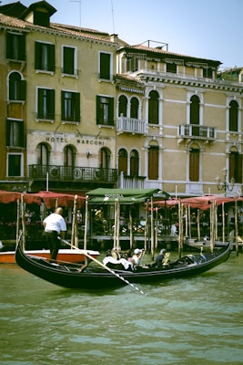 A picturesque canal scene with a gondolier navigating a sleek black gondola through the water. Passengers, leisurely seated under a shaded canopy, enjoy the view of historic, pastel-colored buildings along the water's edge. The architecture features ornate balconies, arched windows, and an aged patina, adding to the charm of the setting.